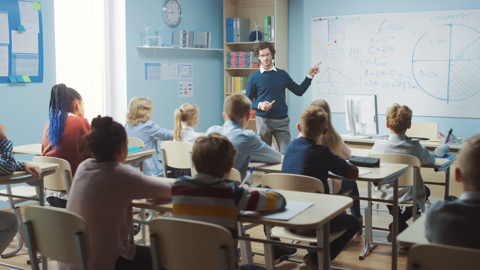 A teacher explaining a lesson at a classroom whiteboard to a class seated at desks