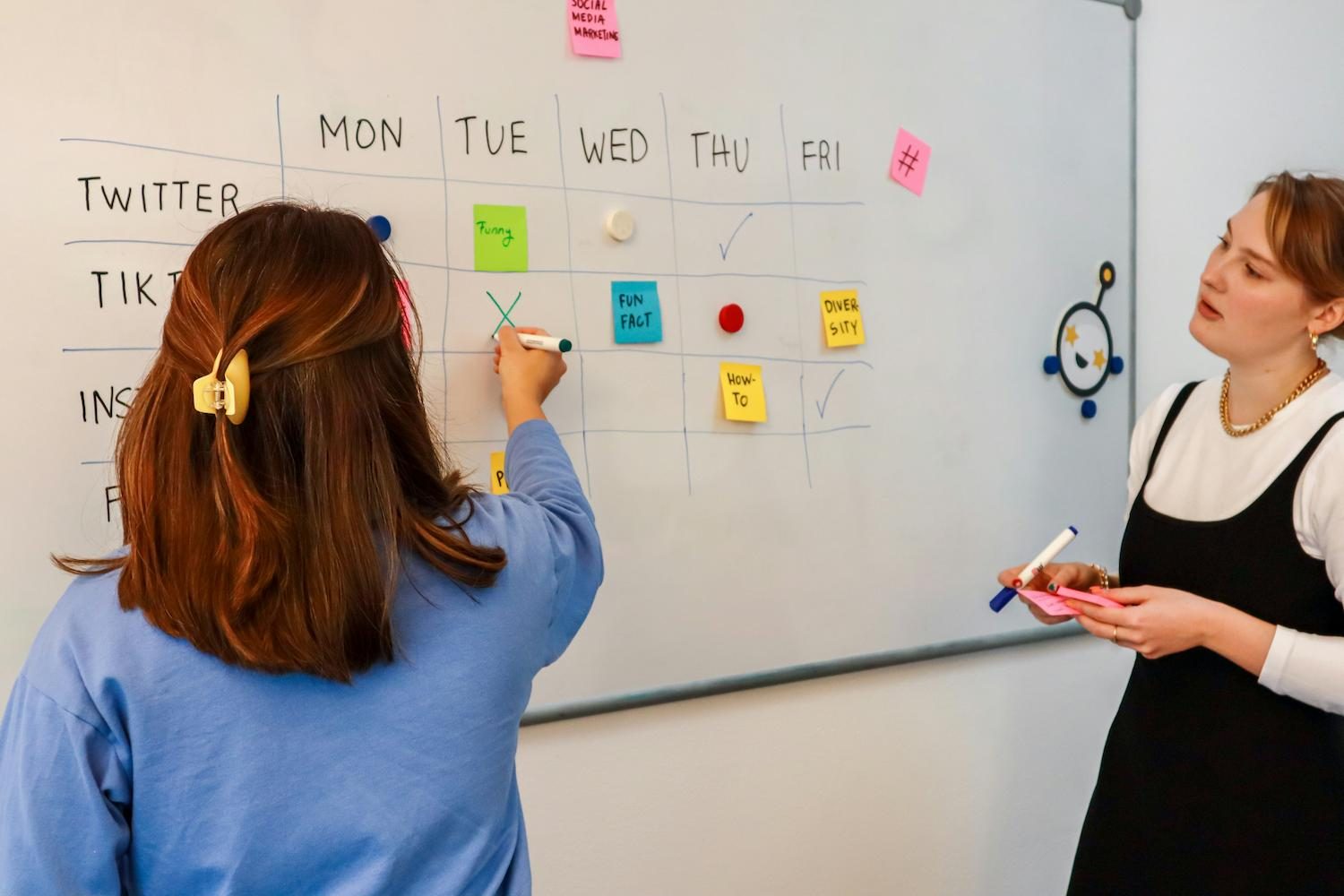 Two colleagues planning a weekly schedule with sticky notes on a whiteboard